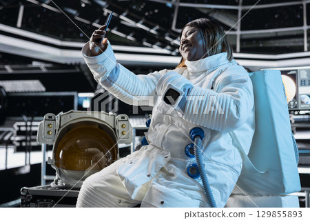 Cheerful female astronaut in starship travelling to space doing selfies using cellphone camera. Happy woman aboard orbit station having fun doing pictures on phone during cosmic journey Cheerful female astronaut in starship travelling to space doing selfies using cellphone camera. Happy woman aboard orbit station having fun doing pictures on phone during cosmic journey 129855893