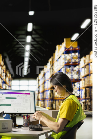 Logistics worker printing and applying awb labels on packages, wearing hi vis vest in a warehouse with barcode airway bill and shipping labels, ensuring order fulfillment and inventory tracking. 129855899