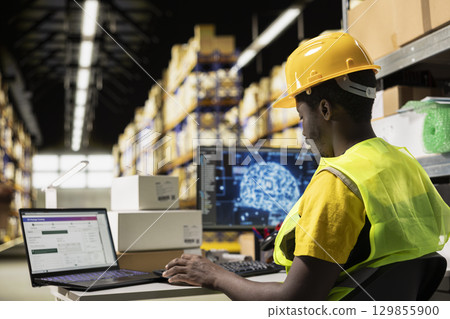 Black employee works with AI tools to enhance package tracking efficiency, using machine learning and parcel tracking info to ensure seamless distribution operations in warehouse. 129855900
