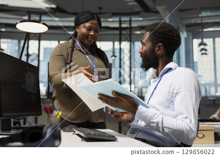 Manager in office handing paperwork to employee working with AI assistant on PC. Worker supervised by team leader while using artificial intelligence to streamline administrative tasks, camera A. 129856022