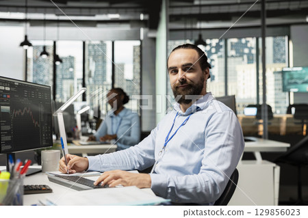 Portrait of smiling trader taking notes while analyzing candlestick charts and price graphs on screen. Cheerful proprietary firm employee writing analysis of market trends and trading strategies Portrait of smiling trader taking notes while analyzing candlestick charts and price graphs on screen. Cheerful proprietary firm employee writing analysis of market trends and trading strategies 129856023