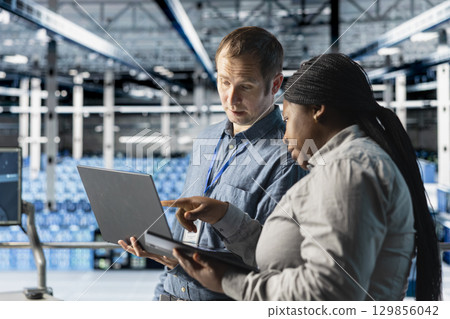 Team of data center admins using laptop to manage software updates for system stability. Teamworking server room IT specialists optimizing performance with device, configuring infrastructure 129856042