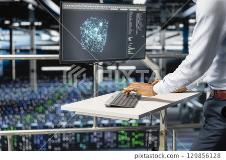 Black software engineer reviews machine learning systems on computer, standing on industrial platform at elevated workstation. Server hub technician monitoring AI infrastructure. 129856128