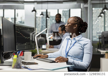 Trader in brokerage firm office analyzing market metrics and executing trades using trading app panel on PC. African american woman working in prop firm monitoring stock exchange 129856131