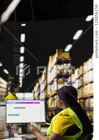 African american female placing adhesive shipping labels on cardboard boxes, preparing the packages for express delivery and applying awb for parcel status tracking on pc software. 129856156