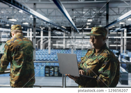 Woman in server room command center managing military operations using laptop. African american soldier wearing uniform running diagnostics on infrastructure in army data center Woman in server room command center managing military operations using laptop. African american soldier wearing uniform running diagnostics on infrastructure in army data center 129856207