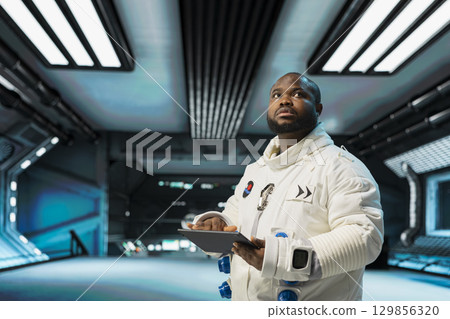 Astronaut in spacecraft verifying navigation coordinates using device. African american man in spaceship updating flight log on tablet during planetary exploration flight Astronaut in spacecraft verifying navigation coordinates using device. African american man in spaceship updating flight log on tablet during planetary exploration flight 129856320