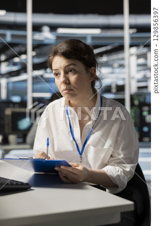 Data center technician taking notes after inspecting gear and checking performance diagnostics. Server farm employee writing repairing needed after conducting system checks Data center technician taking notes after inspecting gear and checking performance diagnostics. Server farm employee writing repairing needed after conducting system checks 129856397