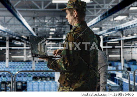 Military officer in data center monitoring warfare drone telemetry on laptop dashboard. IT specialist in army server hub using tactical interface on notebook to support aerial operations 129856400