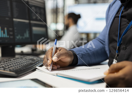 Close up of african american worker writes notes on textbook in the office, analyzing charts and graphs to align company objectives with strategy. Drive progress and measurable performance. 129856414