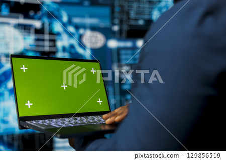 Young male specialist reviews algorithmic data next to green screen, uses laptop surrounded by digital visuals. AI smart technology and neural networks in a high tech environment. 129856519