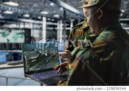Team of army experts in data center reviewing satellite intelligence feeds on laptop. Military units in server farm command center supporting battlefield strategy with orbital imaging analysis Team of army experts in data center reviewing satellite intelligence feeds on laptop. Military units in server farm command center supporting battlefield strategy with orbital imaging analysis 129856544