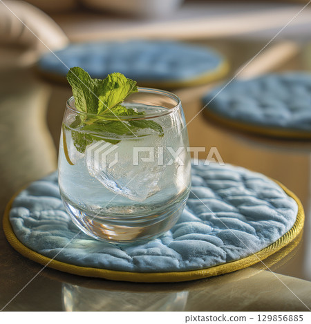 Glass of water with ice and mint on top placed in setting with soft lighting and cozy blue quilted placemats on table, creating refreshing and inviting atmosphere 129856885