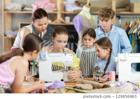 Woman teacher demonstrates how to sew on a sewing machine. Children gather around and watch 129856963