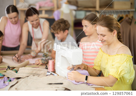 Teenage girl works on sewing machine while teacher and children learn how to cut fabric Teenage girl works on sewing machine while teacher and children learn how to cut fabric 129857061