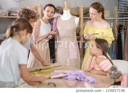 Female dressmaker teacher shows children how to try on dresses on mannequin in classroom 129857146