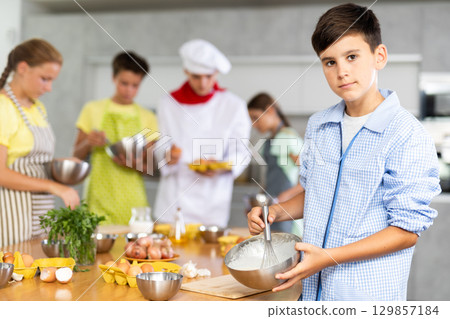Teen boy participant in culinary master class holds dishes with ready-made pancake dough in hands. 129857184