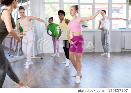 Positive teenage girl practicing ballet positions standing in row with others 129857507