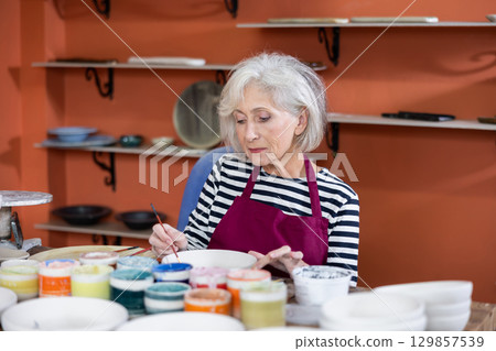 Mature craftswoman painting clay plates acrylic paints in art studio 129857539