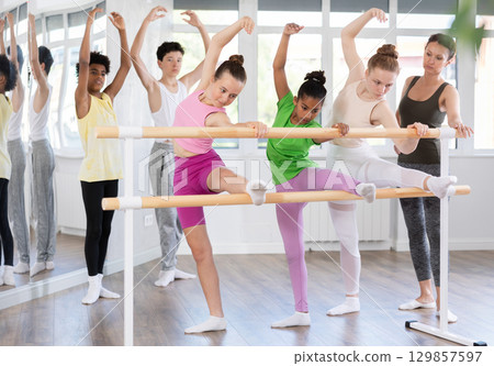 Teenage girls doing leg stretching exercise holding by ballet barre during dance courses 129857597