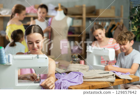 Teenage girl works on sewing machine while teacher and children learn how to cut fabric 129857642