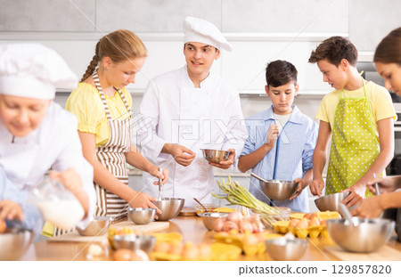 During cooking lesson, male cook tells children about culinary rules, woman assistant helps child During cooking lesson, male cook tells children about culinary rules, woman assistant helps child 129857820