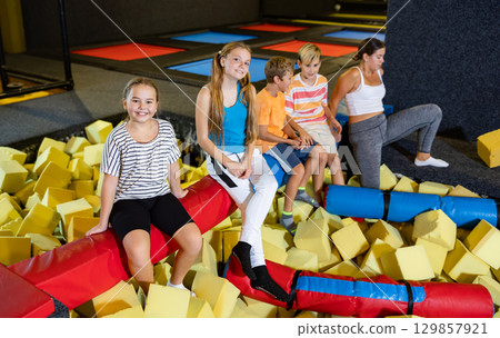 Company of joyful smiling children sitting together on crossbar in entertainment trampoline centre surrounded with foam cubes 129857921