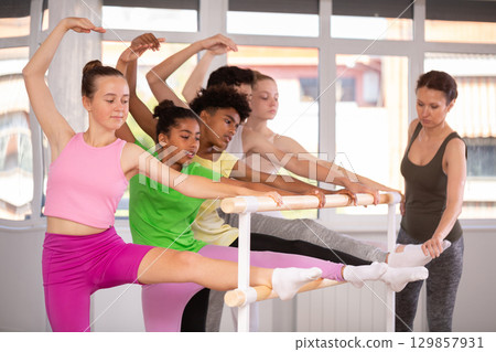 Teenage girl doing leg stretching exercise holding by ballet barre during dance courses 129857931