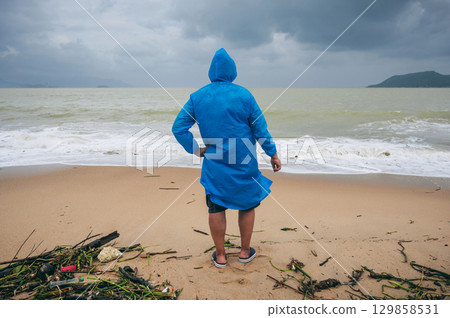 back of man tourist in a raincoat on a dirty beach by the sea after a storm in cloudy weather. The concept of the rainy and monsoon season 129858531
