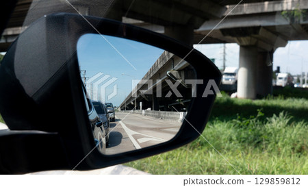 Inside view of mirrors wing. Rear view of a gray car with asphalt road and green trees in the daytime. Clear traffic in rural areas. Grassy area on the side of the road under the concrete overpass. Inside view of mirrors wing. Rear view of a gray car with asphalt road and green trees in the daytime. Clear traffic in rural areas. Grassy area on the side of the road under the concrete overpass. 129859812