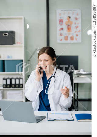 Young female doctor in white medical uniform using laptop and tablet talking video conference call at desk,Doctor sitting at desk Young female doctor in white medical uniform using laptop and tablet talking video conference call at desk,Doctor sitting at desk 129859897