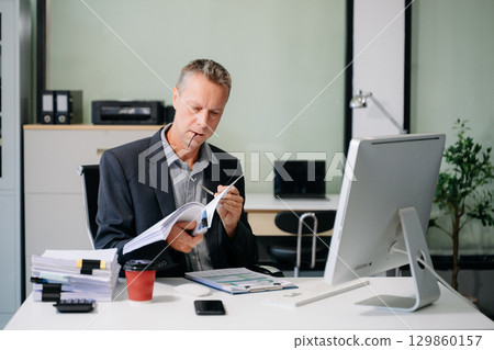 Confident businessman in suit, working on laptop and smartphone at office desk. Professional executive 129860157