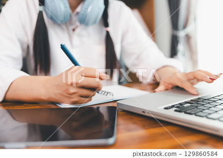 Asian girl student wear headphones learning online watching webinar class looking at laptop computer eLearning remote lesson making notes or video calling virtual conference meeting teacher at home. 129860584