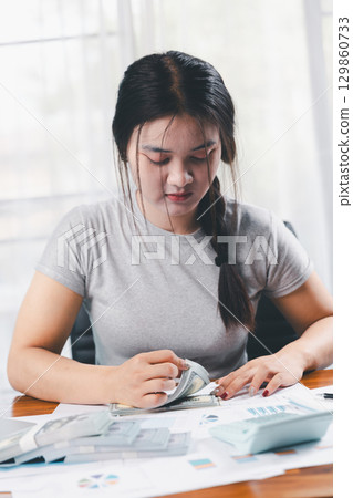 Young Asian woman making stack of coin. invest save finance concept, saving money, investment. dollar bills on table with pile of coins and banks calculator, money to save and invest to make income. 129860733
