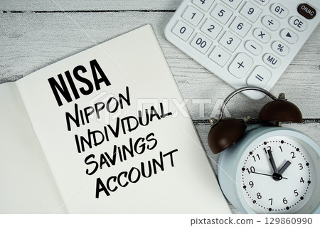 NISA Nippon Individual Savings Account text on paper notebook page with alarm clock and calculator on wooden background 129860990