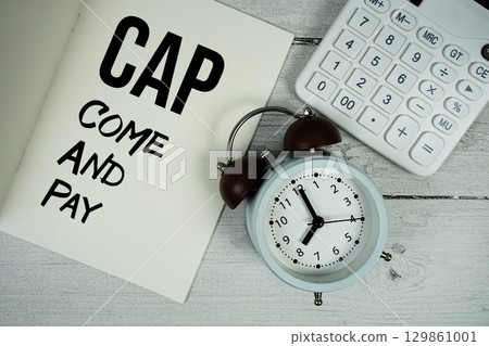 CAP Come And Pay text on paper notebook page with alarm clock and calculator on wooden background 129861001
