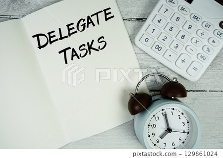 Delegate Tasks text on paper notebook page with alarm clock and calculator on wooden background Delegate Tasks text on paper notebook page with alarm clock and calculator on wooden background 129861024