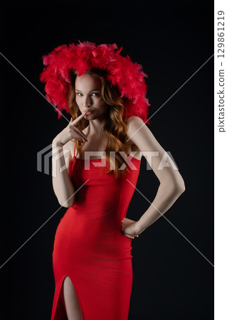 Woman in a striking red dress poses with a playful expression in a studio setting, showcasing her confident allure with a feathered headpiece Woman in a striking red dress poses with a playful expression in a studio setting, showcasing her confident allure with a feathered headpiece 129861219