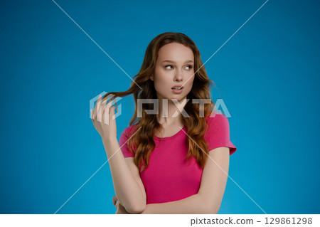 Woman posing in studio with playful expression and vibrant blue background while gently touching her hair 129861298