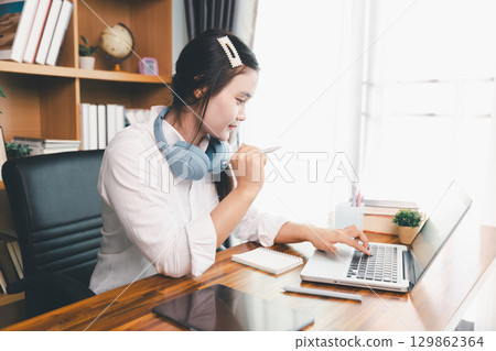 Asian girl student wear headphones learning online watching webinar class looking at laptop computer eLearning remote lesson making notes or video calling virtual conference meeting teacher at home. Asian girl student wear headphones learning online watching webinar class looking at laptop computer eLearning remote lesson making notes or video calling virtual conference meeting teacher at home. 129862364