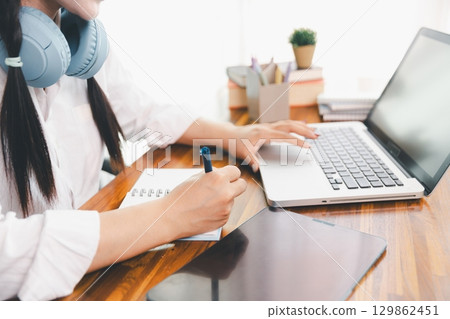 Asian girl student wear headphones learning online watching webinar class looking at laptop computer eLearning remote lesson making notes or video calling virtual conference meeting teacher at home. 129862451