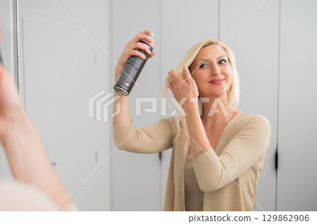 Elegant blonde woman in nude outfit applying hair spray to style and fix her hairstyle while standing by mirror indoors 129862906