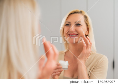 Elegant blonde woman in nude outfit using moisturizing cream to soften and hydrate skin while looking in mirror indoors Elegant blonde woman in nude outfit using moisturizing cream to soften and hydrate skin while looking in mirror indoors 129862907