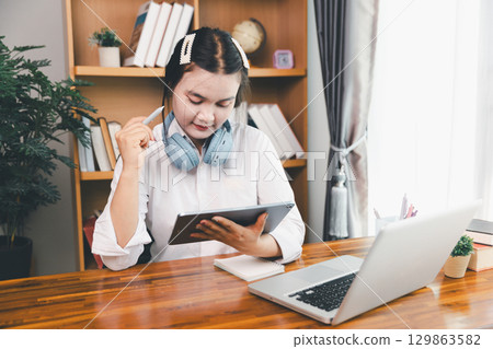Asian girl student wear headphones learning online watching webinar class looking at laptop computer eLearning remote lesson making notes or video calling virtual conference meeting teacher at home. 129863582