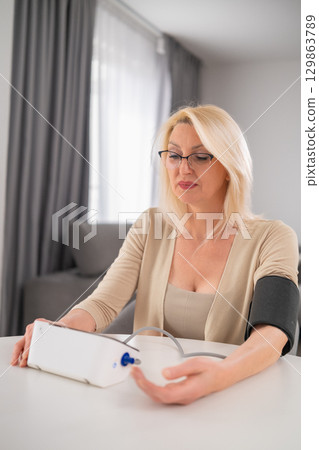 Blonde middle aged woman in beige outfit measuring blood pressure at home in modern apartment living room Blonde middle aged woman in beige outfit measuring blood pressure at home in modern apartment living room 129863789