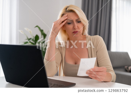 Blonde woman in beige outfit sitting at home, looking upset at a bill and struggling with financial difficulties, economic stress concept 129863792