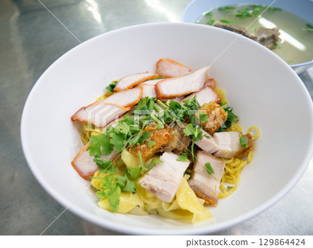 Chinese egg noodles with roasted red pork, crispy pork belly, and wontons in a bowl on table. Chinese egg noodles with roasted red pork, crispy pork belly, and wontons in a bowl on table. 129864424