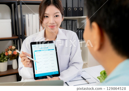 Asian nutritionist using tablet to explaining personalized meal plan with AI to patient. Asian nutritionist using tablet to explaining personalized meal plan with AI to patient. 129864528