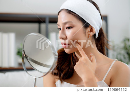 Young asian woman looking mirror with stress on facial sitting on sofa in living room at home. 129864532