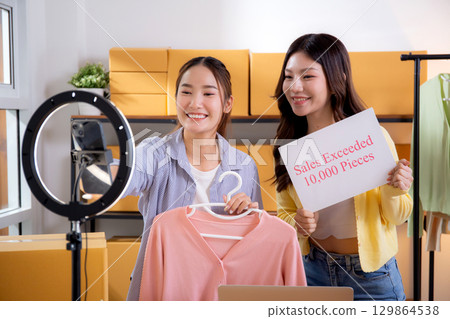 Two asian woman live streaming online clothing store while showing clothing with smartphone. 129864538